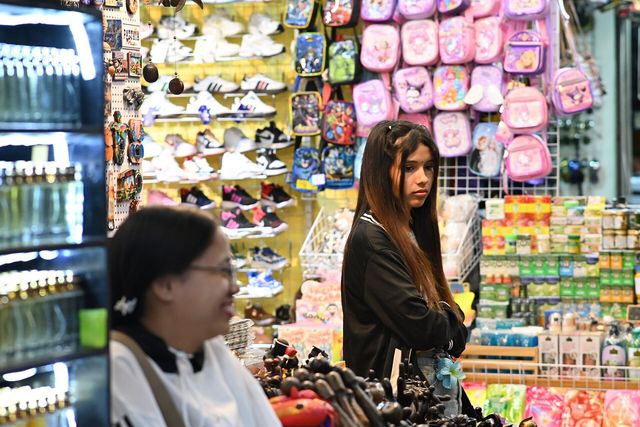 DFC 3749 A young woman browses a busy market stall filled with colorful backpacks shoes and packaged goods whi