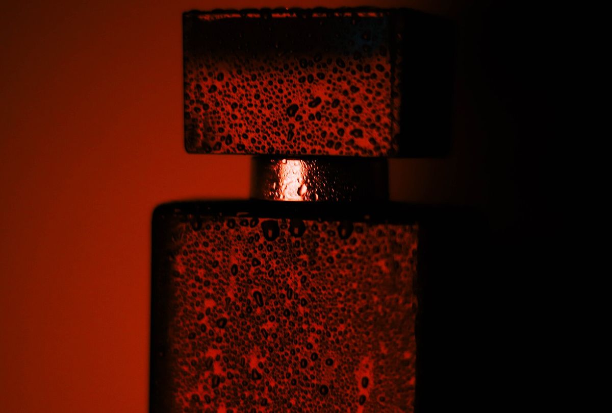 A close-up artistic photograph of a perfume bottle covered in water droplets, illuminated by dramatic red and